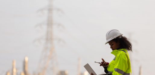 A female engineer in a high-visibility jacket and helmet uses a laptop and smartphone near a power plant and transmission towers, illustrating modern technology, communication, and industrial work